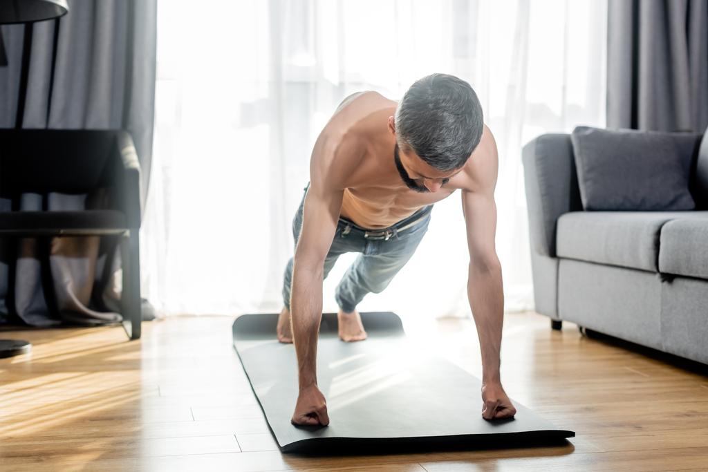 Shirtless man doing press ups while training on fitness mat in living room - Photo, Image