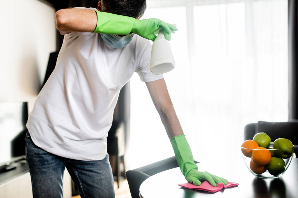 Uomo in maschera medica e guanti di gomma tavolo di pulizia con straccio e detergente a casa
  - Foto, immagini