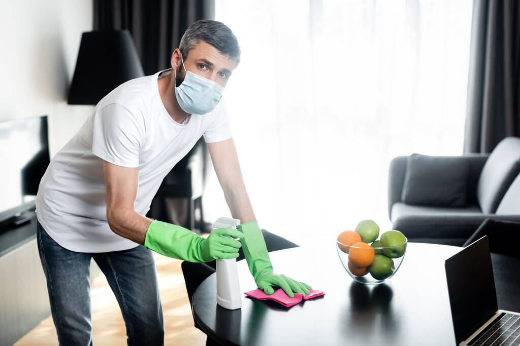 Άντρας με ιατρική μάσκα και γάντια από καουτσούκ που κρατούν κουρέλια και απορρυπαντικά στο σαλόνι - Φωτογραφία, εικόνα