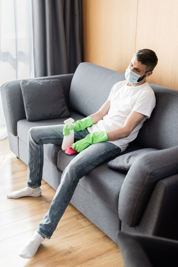 Selektiver Fokus des Mannes in medizinischer Maske, der Lappen und Waschmittel hält, während er auf der Couch sitzt - Foto, Bild