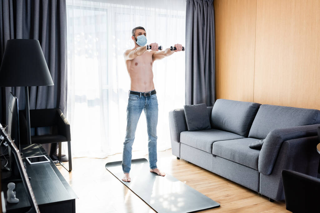 Muž bez trička v lékařské masce trénink s činkami na fitness podložky v obývacím pokoji - Fotografie, Obrázek