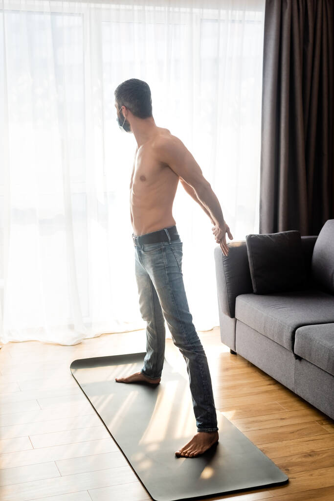 Widok z boku człowieka w masce medycznej rozciągającej się na macie fitness w domu  - Zdjęcie, obraz
