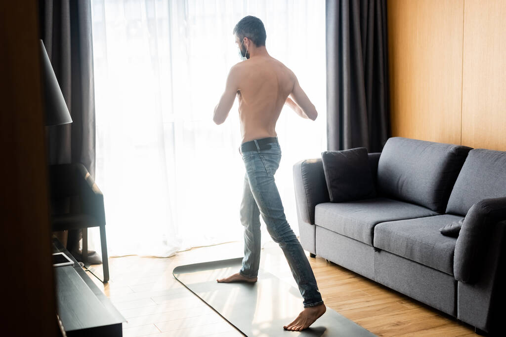 Muž bez trička v lékařské masce trénink na fitness podložky v obývacím pokoji - Fotografie, Obrázek