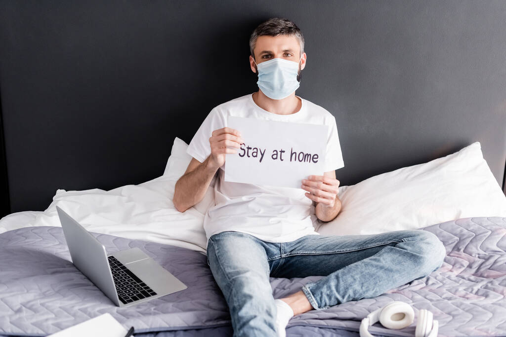 Man in medical mask holding card with stay at home lettering near laptop and headphones on bed  - Photo, Image