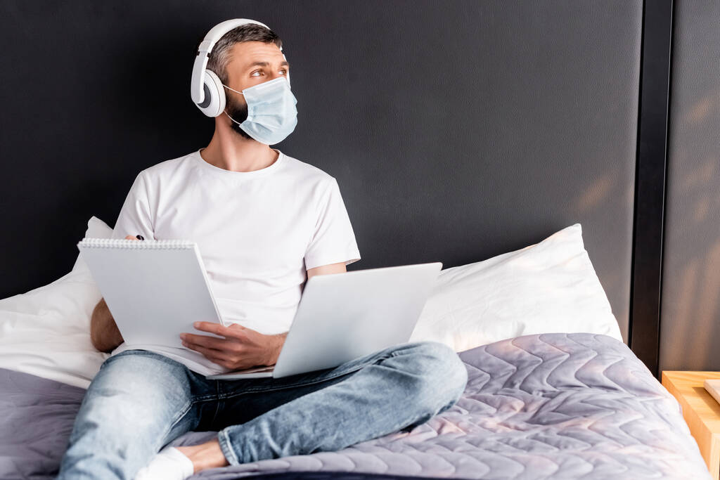 Freelancer en máscara médica y auriculares que trabajan con portátil y portátil en la cama
  - Foto, Imagen