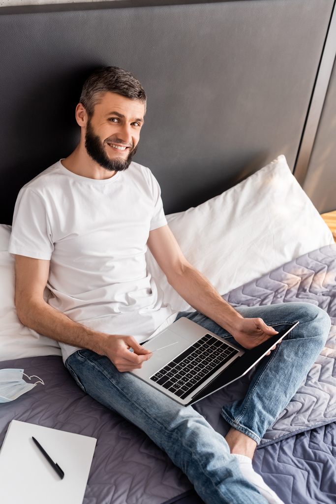 Vista ad alto angolo di uomo sorridente in possesso di carta di credito e laptop vicino alla maschera medica e notebook sul letto
  - Foto, immagini