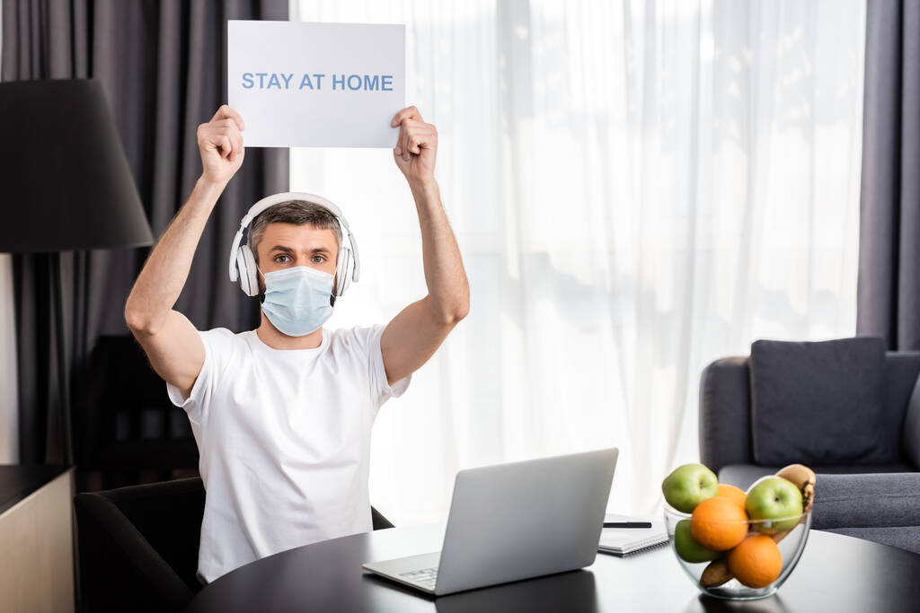 Freelancer en máscara médica y auriculares con tarjeta de retención con letras en casa cerca de la computadora portátil y frutas en la mesa
  - Foto, Imagen