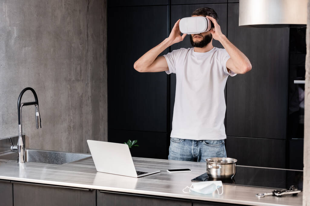 Άνθρωπος που χρησιμοποιεί ακουστικά εικονικής πραγματικότητας κοντά σε ψηφιακές συσκευές και ιατρική μάσκα σε πάγκο κουζίνας  - Φωτογραφία, εικόνα