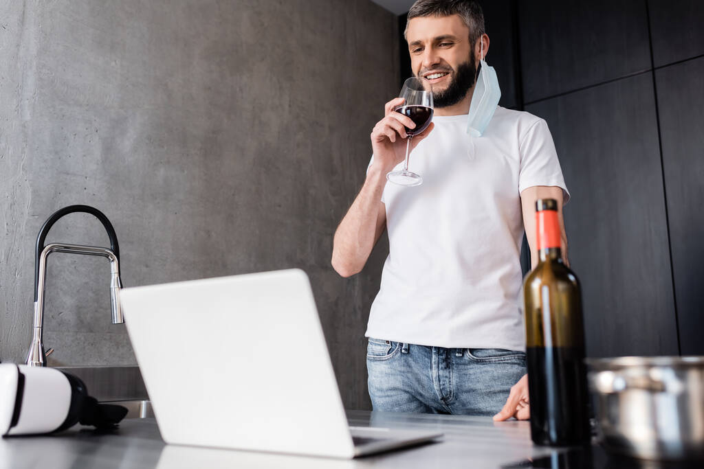 Focus selettivo di freelance sorridente in maschera medica bere vino vicino al computer portatile sul piano di lavoro in cucina
  - Foto, immagini