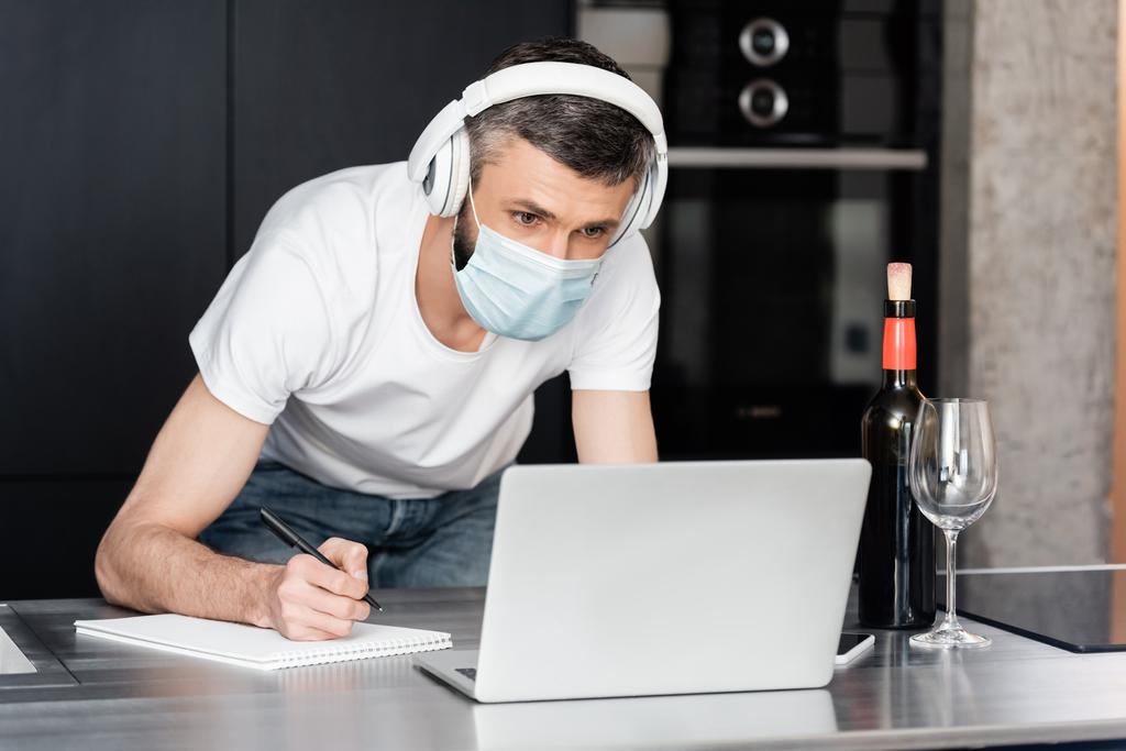 Teleworker in headphones and medical mask writing on notebook near laptop and wine on worktop in kitchen  - Photo, Image