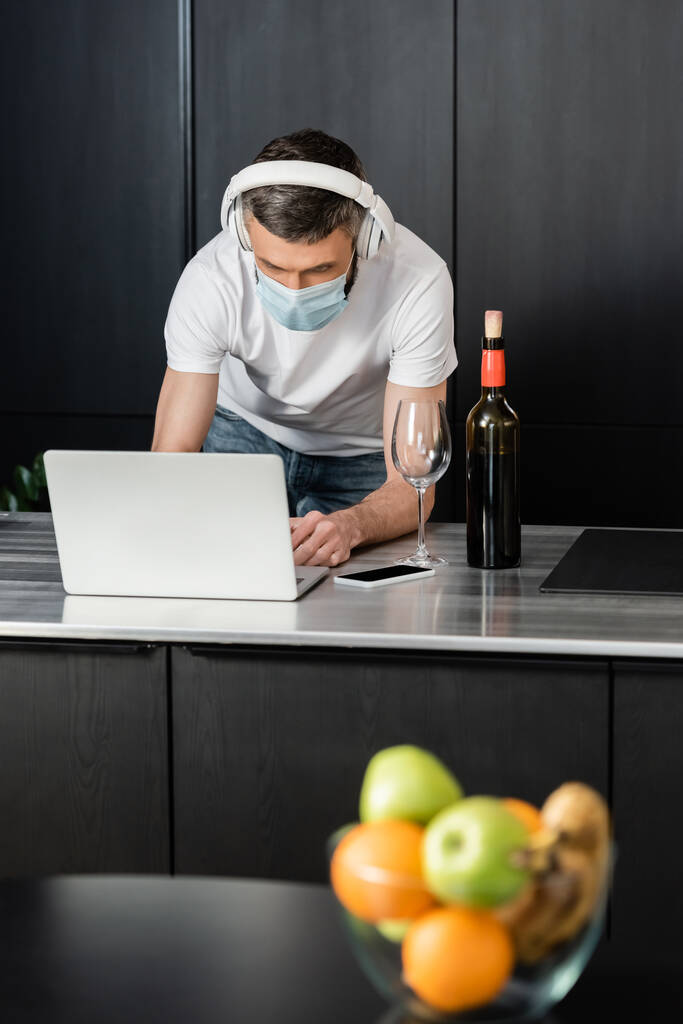 Enfoque selectivo del freelancer en auriculares y máscara médica usando portátil cerca del vino en la encimera de la cocina
  - Foto, imagen