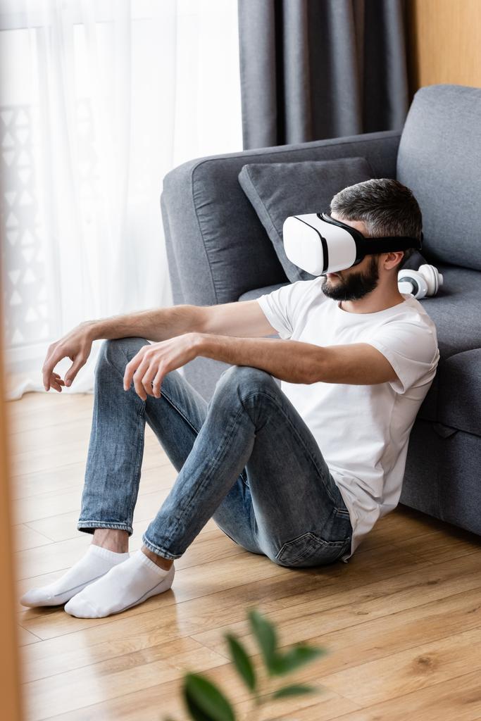 VR kulaklıklı adamın seçici odak noktası yerde, kanepenin yanında oturması.  - Fotoğraf, Görsel
