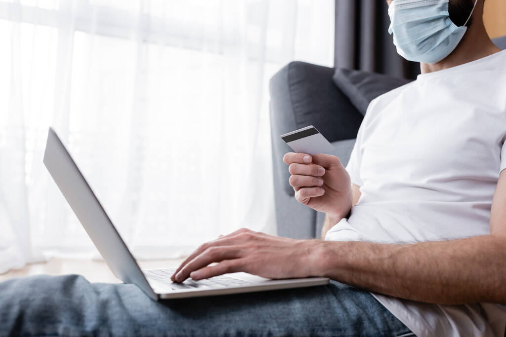 Gewassen beeld van de man met medisch masker met behulp van laptop en creditcard thuis  - Foto, afbeelding
