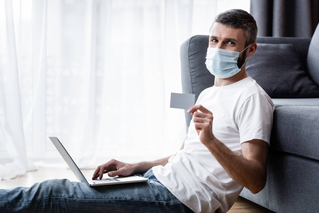 Vista lateral do homem em máscara médica olhando para a câmera enquanto segurando cartão de crédito e usando laptop em casa
 - Foto, Imagem