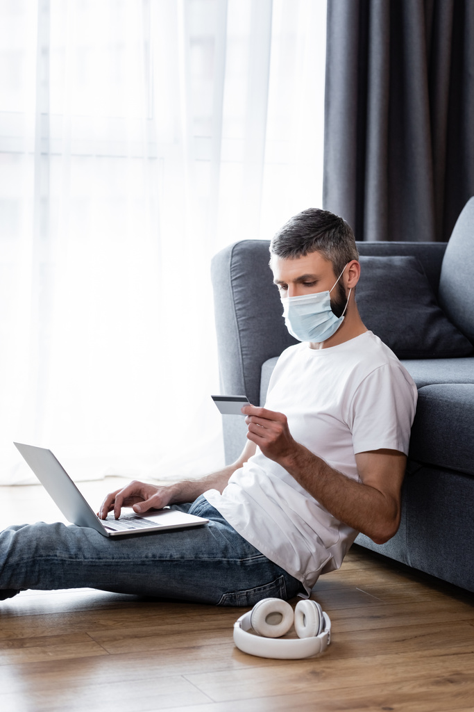 Vista laterale dell'uomo in maschera medica utilizzando laptop e carta di credito vicino alle cuffie sul pavimento in soggiorno
 - Foto, immagini