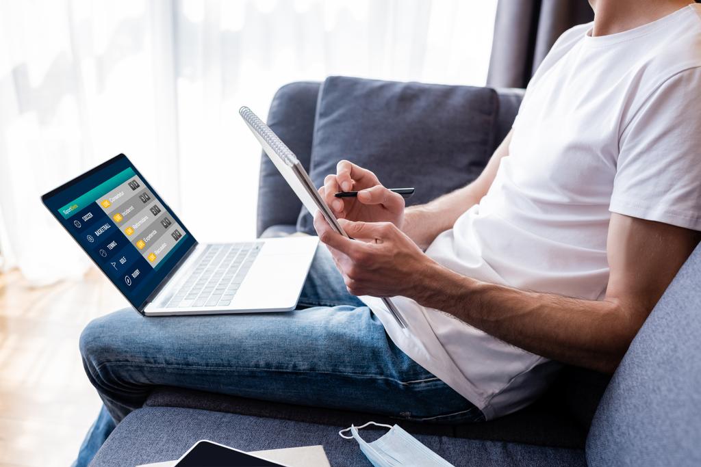 Обрезанный вид человека, пишущего на ноутбуке рядом с ноутбуком со спортивными ставками на экране и медицинской маской на диване
  - Фото, изображение