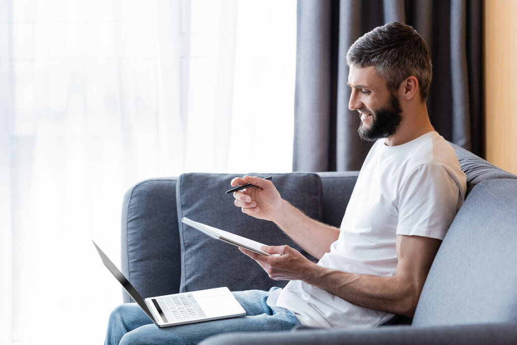 Side view of smiling freelancer holding pen and notebook near laptop on sofa at home  - Photo, Image