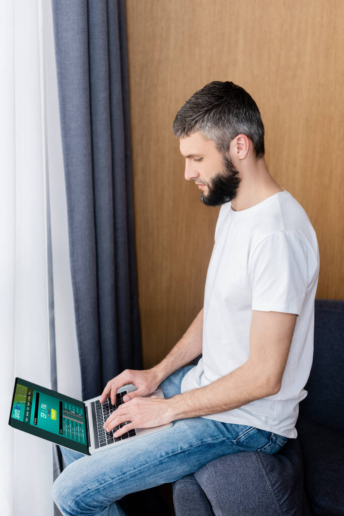 Вид сбоку на человека, использующего ноутбук со спортивным сайтом на диване
  - Фото, изображение
