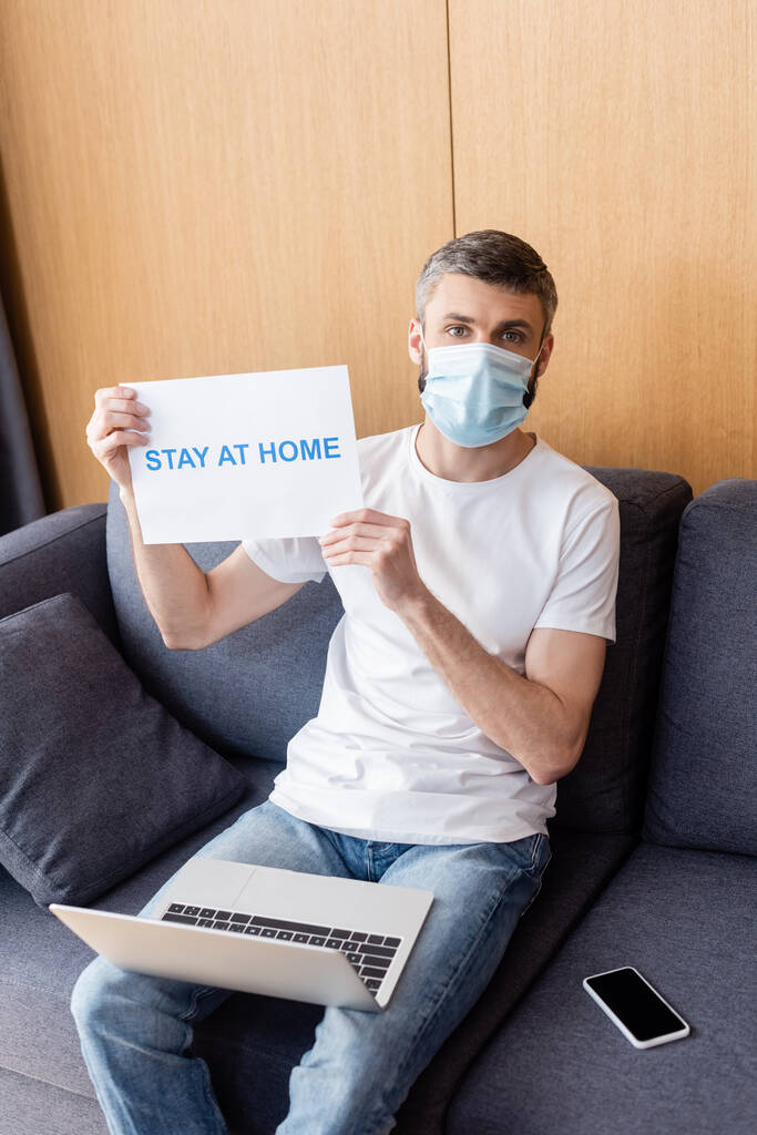 Freelancer w masce medycznej trzymając kartę z pobytu w domu litery w pobliżu urządzeń cyfrowych na kanapie  - Zdjęcie, obraz