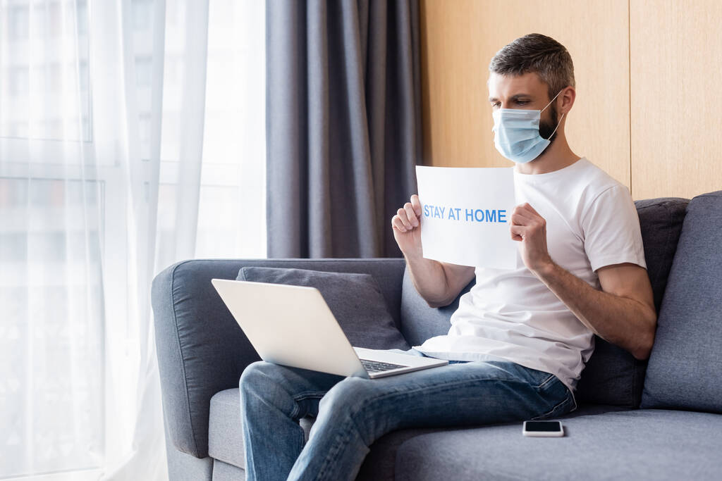 Man in medical mask holding card with stay at home lettering ενώ κοιτάς το laptop στον καναπέ  - Φωτογραφία, εικόνα