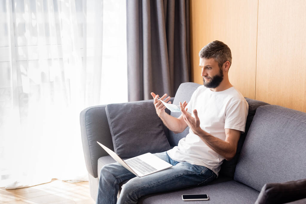 Uomo con maschera medica vicino a dispositivi digitali sul divano in soggiorno
 - Foto, immagini