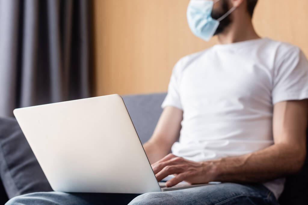 Selektywne skupienie freelancer w masce medycznej przy użyciu laptopa na sofie w domu  - Zdjęcie, obraz