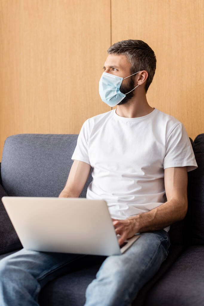 Επιλεκτική εστίαση του τηλεργαζόμενου με ιατρική μάσκα κοιτάζοντας αλλού ενώ χρησιμοποιεί φορητό υπολογιστή στο σπίτι  - Φωτογραφία, εικόνα