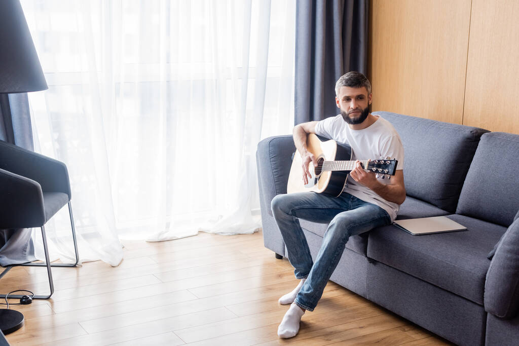 Bonito homem tocando guitarra acústica perto de laptop no sofá em casa
  - Foto, Imagem