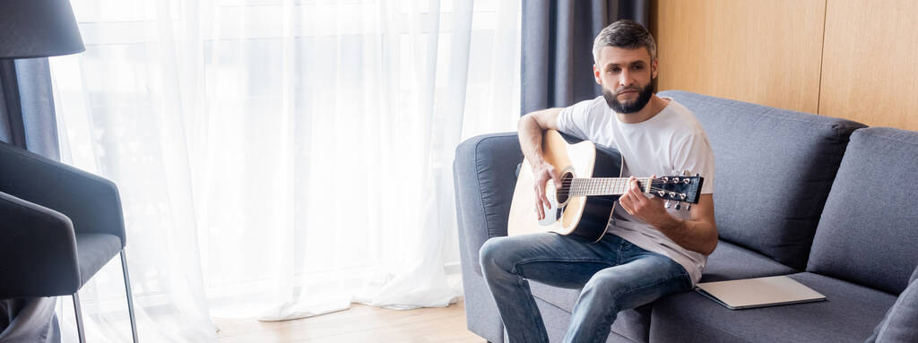 Panoramische opname van een knappe man die naar de camera kijkt terwijl hij akoestische gitaar speelt op de bank in de woonkamer - Foto, afbeelding