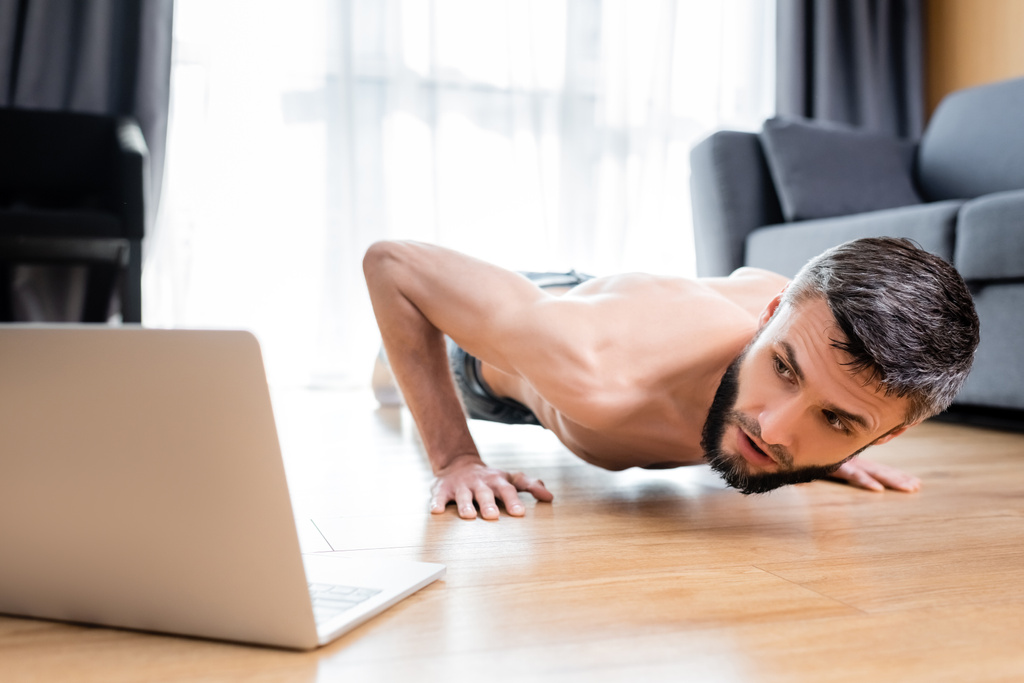 Selektywne skupienie sportowca bez koszulki robi pompki w pobliżu laptopa na podłodze  - Zdjęcie, obraz