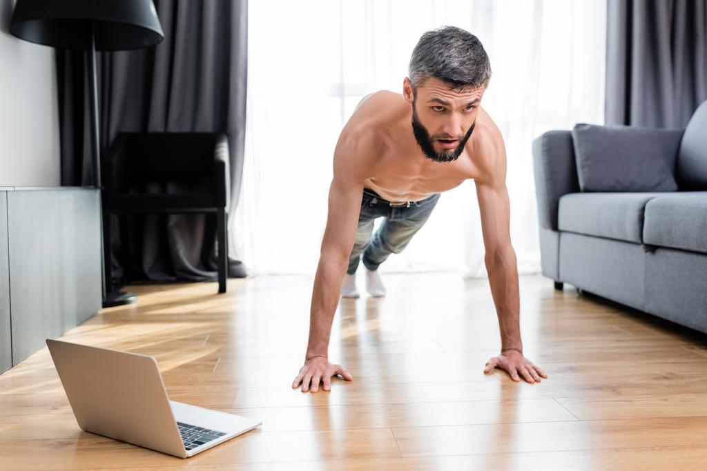 Homem muscular fazendo press ups no chão perto de laptop na sala de estar
 - Foto, Imagem