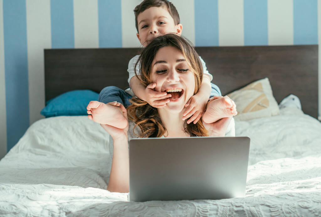 maluch chłopiec dotykając wesoły freelancer matka w pobliżu laptopa na łóżku  - Zdjęcie, obraz