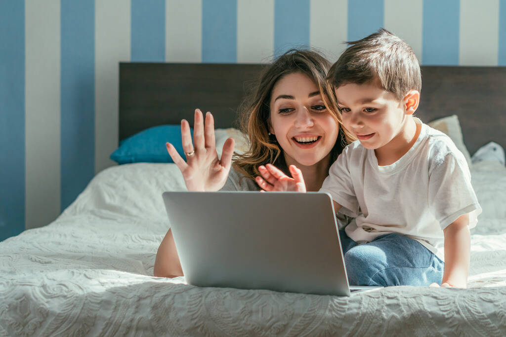 szczęśliwy matka i mały chłopiec macha rękami podczas mając wideo połączenie w sypialni  - Zdjęcie, obraz
