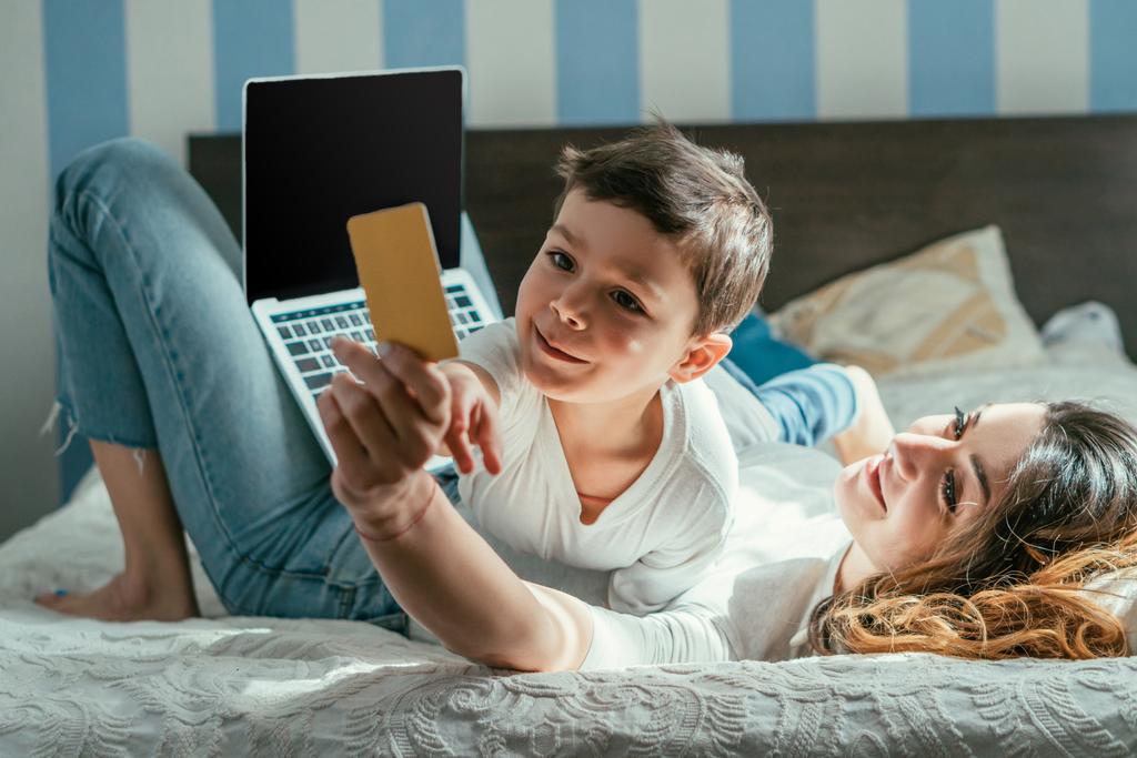 enfoque selectivo de la mujer acostada en la cama y la celebración de la tarjeta de crédito cerca de la computadora portátil con pantalla en blanco y lindo niño pequeño
 - Foto, imagen