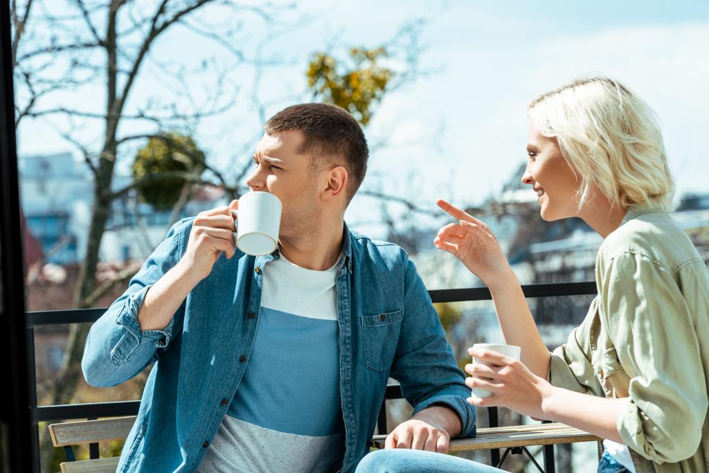 smiling couple talking and drinking tea on terrace while woman pointing with finger away - Photo, Image