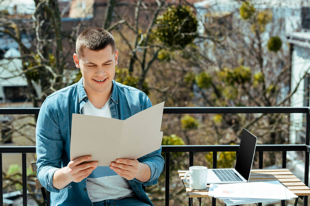 freelance sorridente seduto sulla terrazza con cartella vicino a laptop e carte
 - Foto, immagini