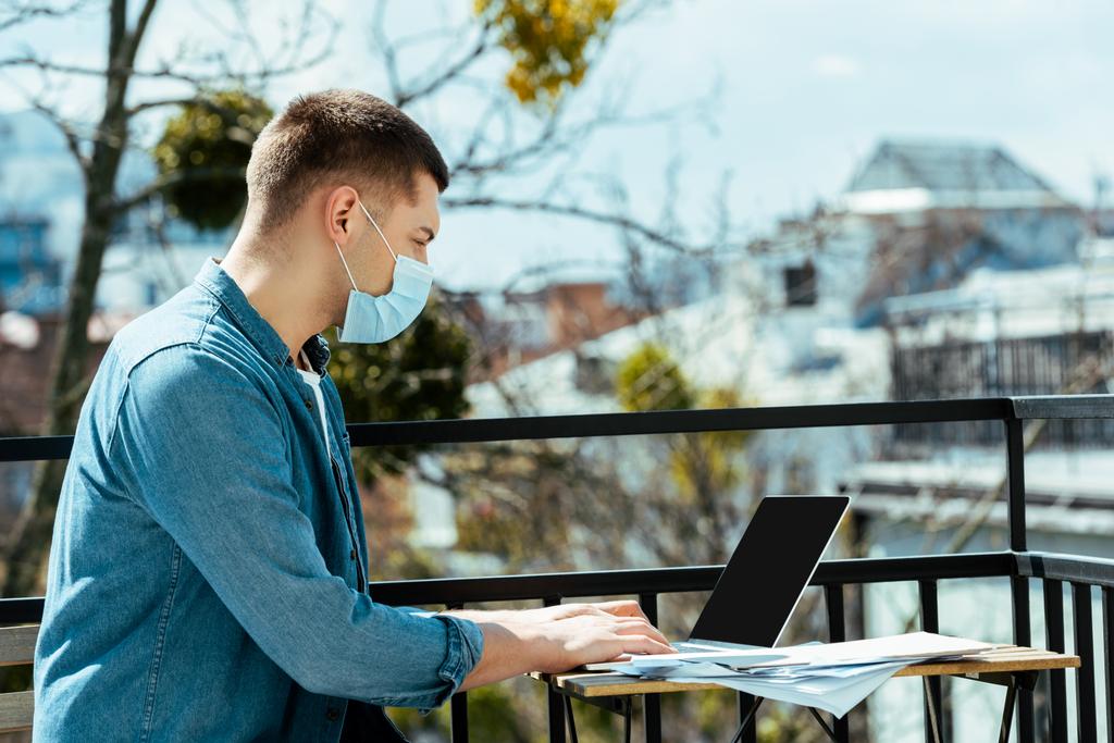 vista lateral del freelancer en máscara médica sentado en la terraza y utilizando el ordenador portátil
 - Foto, Imagen