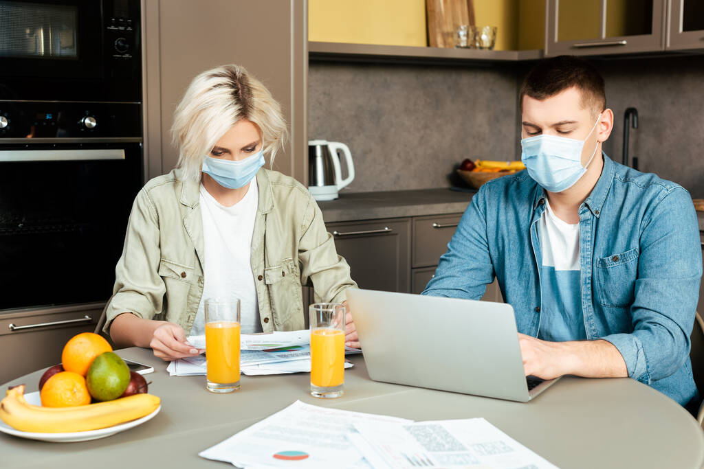 para w maskach medycznych pracujących z papierami i laptopem w pobliżu soku pomarańczowego w gazach w kuchni w domu - Zdjęcie, obraz