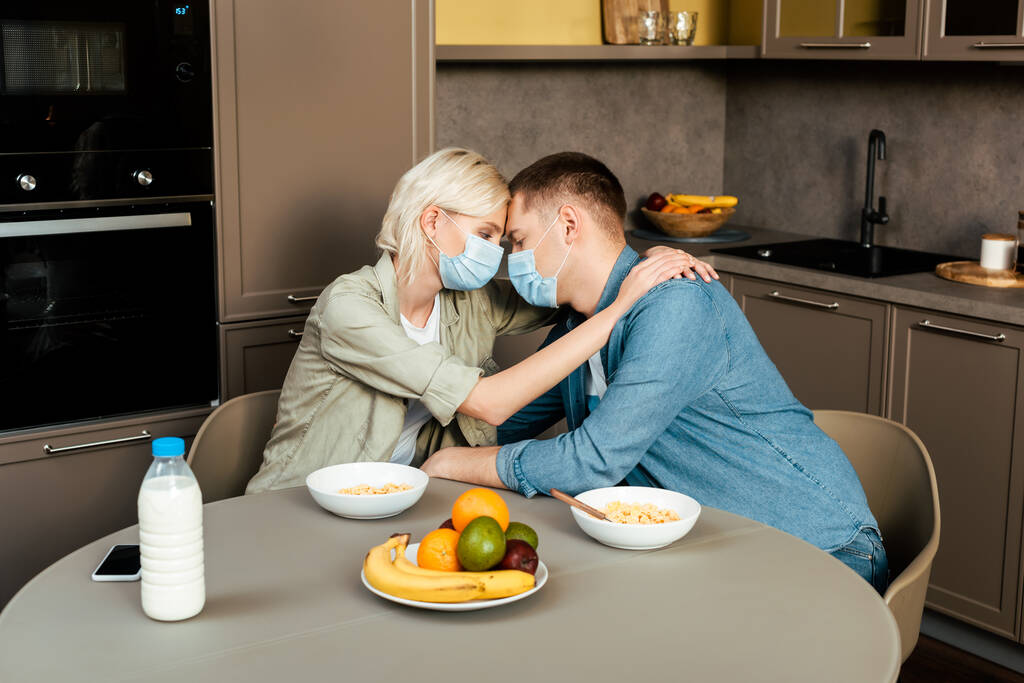 Paar in medizinischen Masken umarmt sich beim Frühstück in der Küche - Foto, Bild