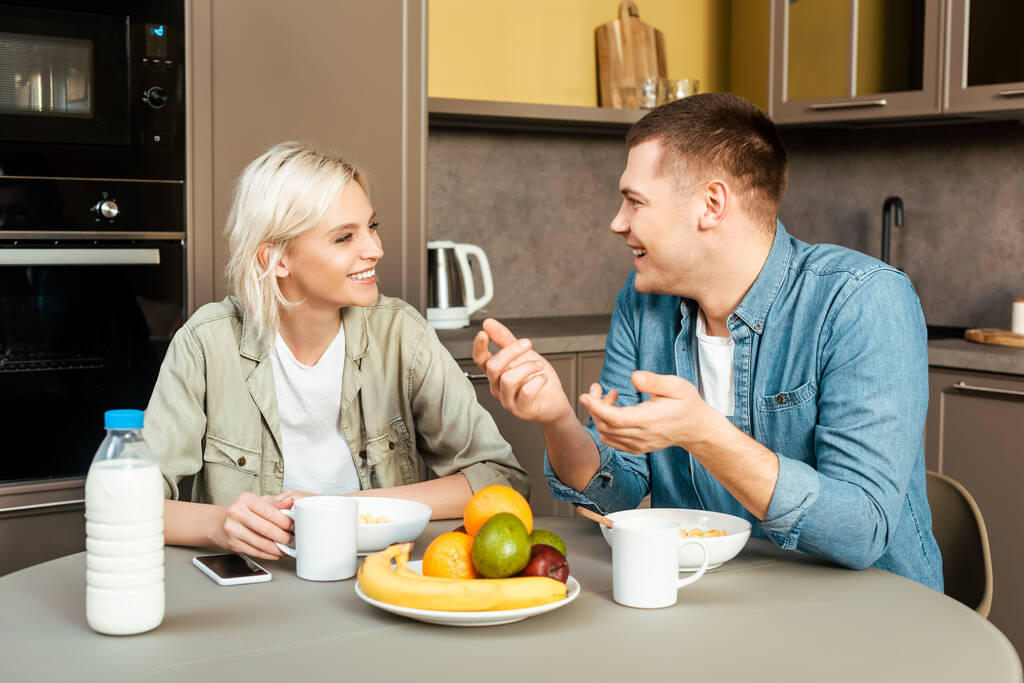 hymyilevä pari puhuu kun ottaa aamiaisen keittiössä
 - Valokuva, kuva