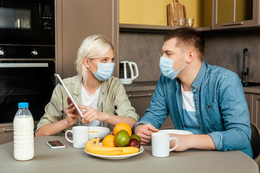 γυναίκα με ιατρική μάσκα που δείχνει ψηφιακή ταμπλέτα στο σύζυγο, ενώ το πρωινό στην κουζίνα - Φωτογραφία, εικόνα