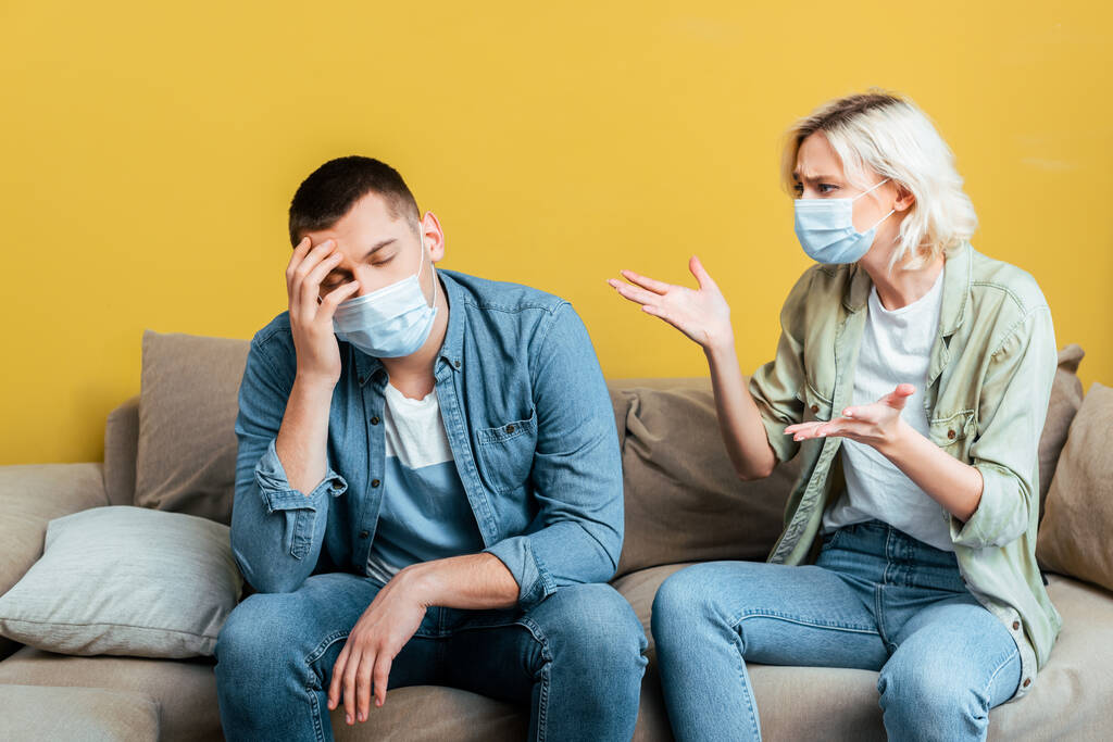 молода пара в медичних масках сваряться на дивані біля жовтої стіни
 - Фото, зображення