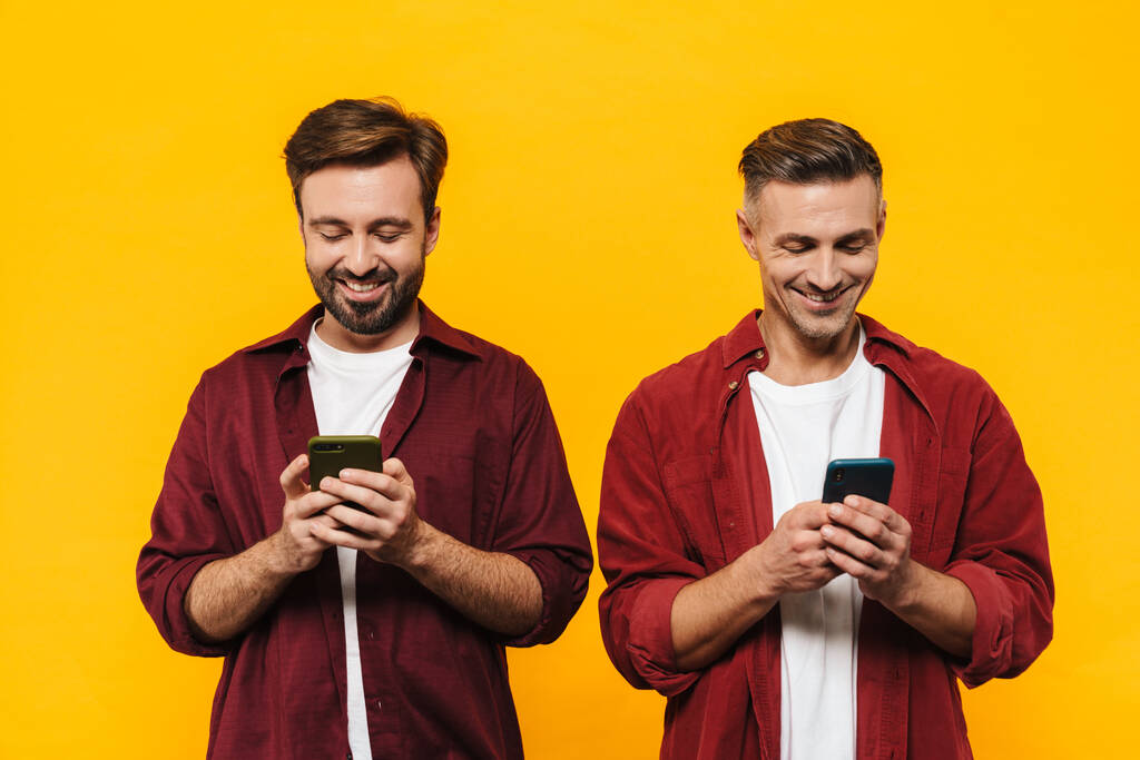 Imagen de dos hombres guapos de 30 años en camisas rojas sonriendo y manteniendo juntos los teléfonos celulares aislados sobre fondo amarillo
 - Foto, Imagen
