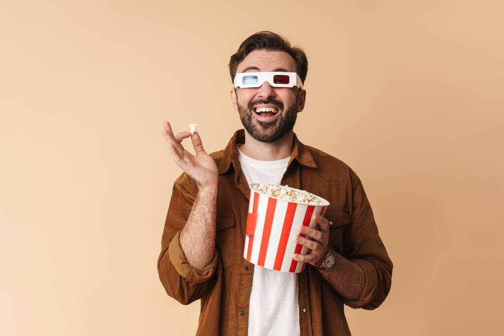 Portrait of a cheerful young arttractive bearded man wearing casual clothes standing isolated over beige background, watching a movie in 3d glasses, eating popcorn - Photo, Image
