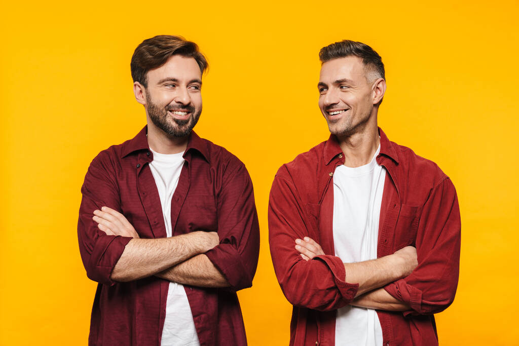 Imagen de dos hombres guapos de 30 años en camisas rojas sonriendo y de pie con los brazos cruzados aislados sobre fondo amarillo
 - Foto, Imagen
