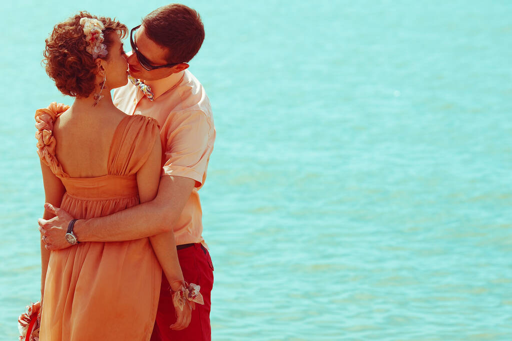 Feliz luna de miel (vacaciones) concepto. Joven feliz casado hipsters en ropa de moda de pie y besándose sobre fondo azul océano (mar). Día soleado de verano. Copiado. tiro al aire libre
 - Foto, Imagen