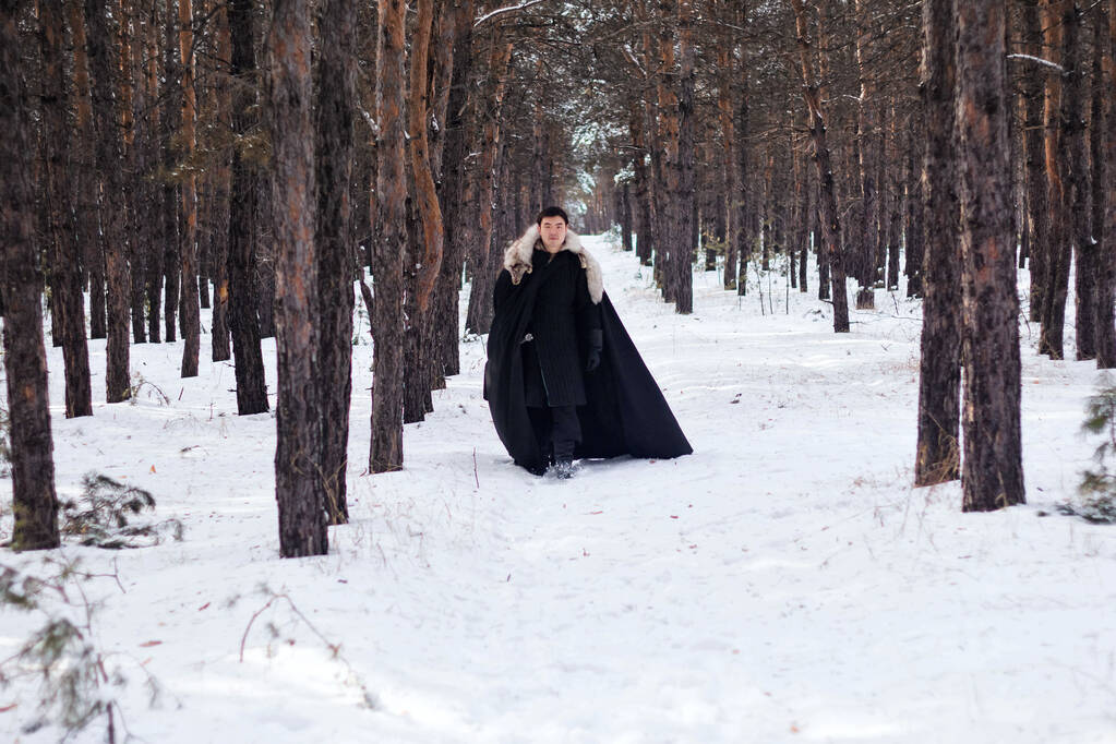 Воїн в чорному стьобаному одязі в чорному плащі з білим хутряним коміром і довгим мечем в руках ходить по дорозі в лісі
. - Фото, зображення