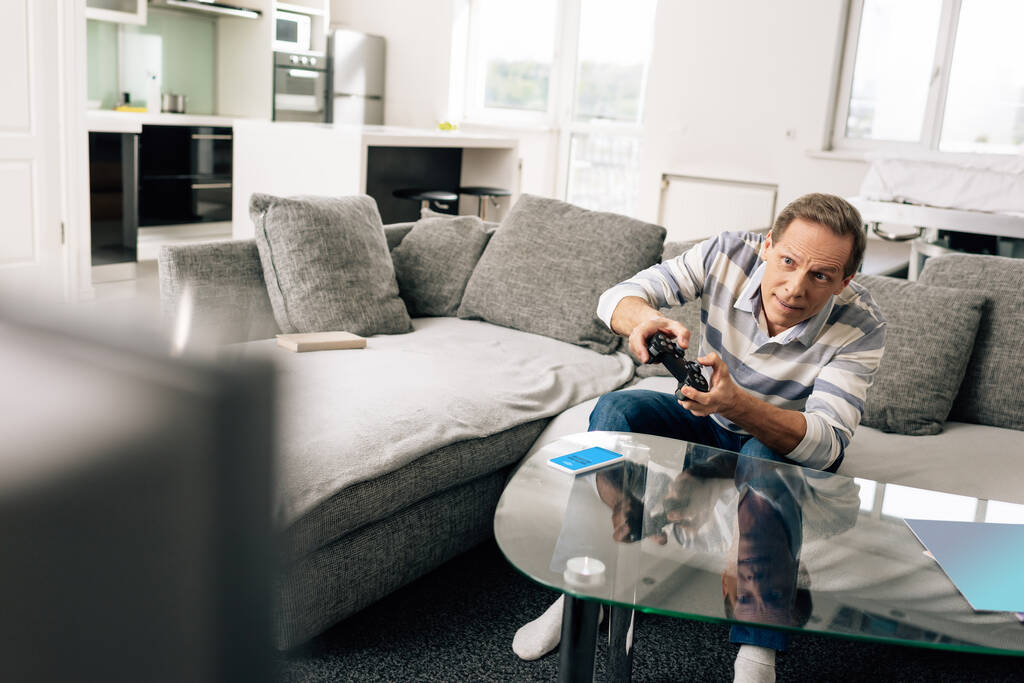 KYIV, UKRAINE - APRIL 14, 2020: selective focus of man playing video game and holding joystick near smartphone with skype app on coffee table  - Photo, Image