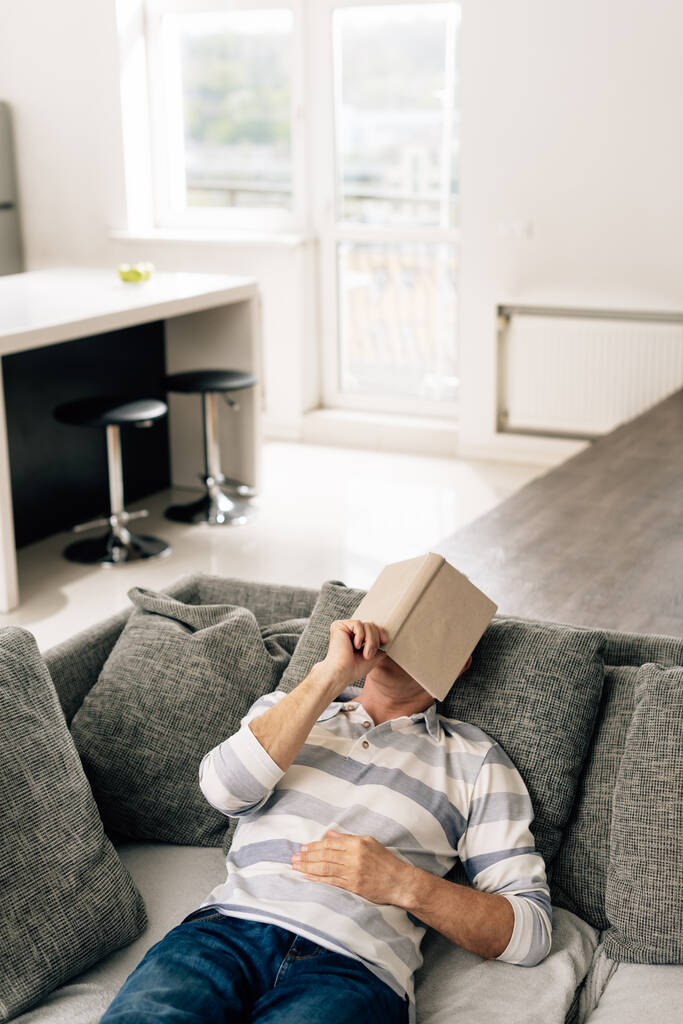 homem deitado no sofá e tocando livro enquanto cobria o rosto na sala de estar
  - Foto, Imagem
