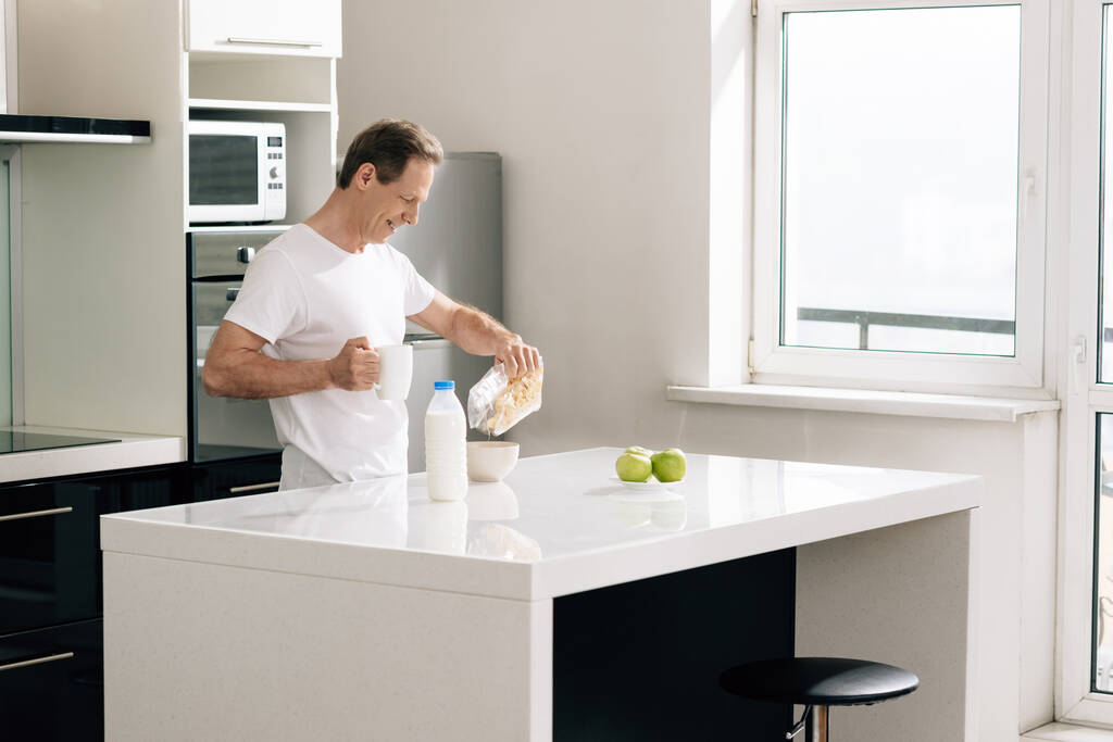 happy man pouring cornflakes in bowl near bottle with milk and apples - Photo, Image